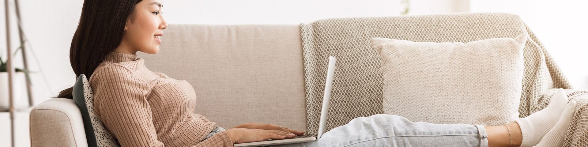 A woman in jeans and a sweater, lies on a beige couch, using a laptop.